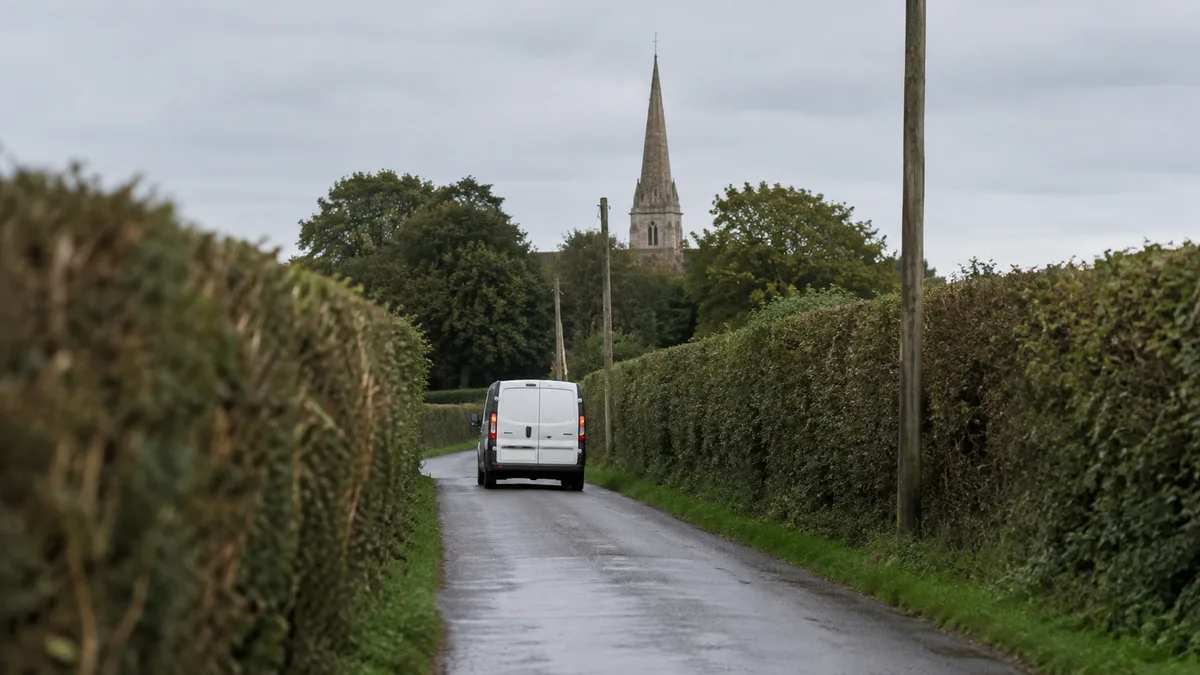 A damp narrow country lane between high hedgerows on an overcast day, with a plain unbranded white van driving away in the middle distance, wooden telegraph poles along the verge and a stone church spire rising over the treetops ahead
