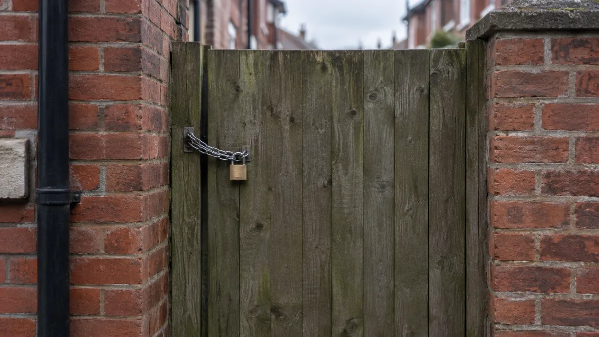 A padlocked wooden side gate on a red brick terrace, secured with a plain unbranded padlock and chain, photographed from the pavement
